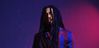 Skip Marley poses in a bold striped suit on a purple and red background to promote the release of his new single “In Our Sight” and official video, filmed in Jamaica.